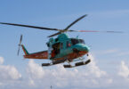 Turquoise rescue helicopter hovering above a sandy beach with rescue crew on the shore and waves in the background, indicating a rescue operation.