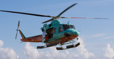 Turquoise rescue helicopter hovering above a sandy beach with rescue crew on the shore and waves in the background, indicating a rescue operation.