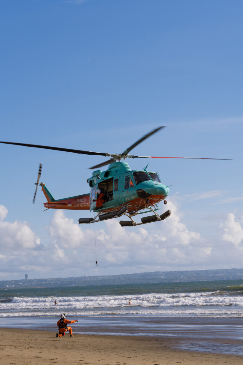 Turquoise rescue helicopter hovering above a sandy beach with rescue crew on the shore and waves in the background, indicating a rescue operation.