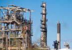 Industrial refinery with metal framework, pipes, and tall smokestacks against a blue sky.