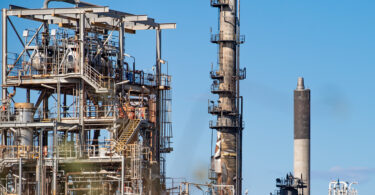Industrial refinery with metal framework, pipes, and tall smokestacks against a blue sky.