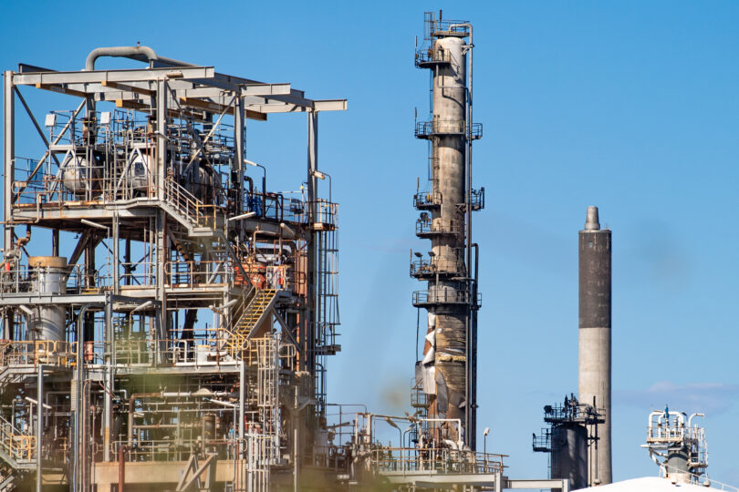 Industrial refinery with metal framework, pipes, and tall smokestacks against a blue sky.
