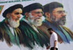 Mural of three bearded men in green robes and turbans on a white wall; a man in a white shirt walks by in the foreground.