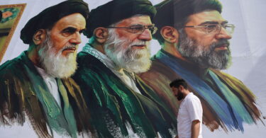 Mural of three bearded men in green robes and turbans on a white wall; a man in a white shirt walks by in the foreground.