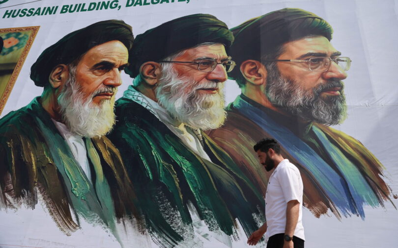 Mural of three bearded men in green robes and turbans on a white wall; a man in a white shirt walks by in the foreground.