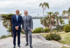 Two men in suits standing together outdoors by the water, with palm trees and a city skyline in the background.