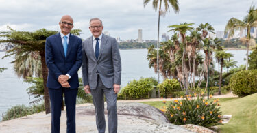 Two men in suits standing together outdoors by the water, with palm trees and a city skyline in the background.