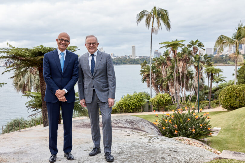Two men in suits standing together outdoors by the water, with palm trees and a city skyline in the background.