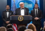 President Donald Trump speaks at a podium flanked by two men in suits during a formal press briefing, with the presidential seal on the lectern and American flags in the background.