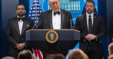 President Donald Trump speaks at a podium flanked by two men in suits during a formal press briefing, with the presidential seal on the lectern and American flags in the background.
