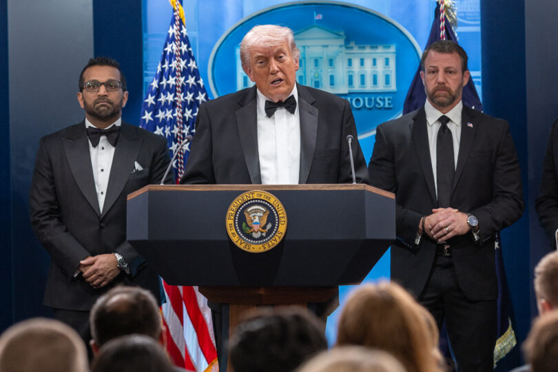 President Donald Trump speaks at a podium flanked by two men in suits during a formal press briefing, with the presidential seal on the lectern and American flags in the background.