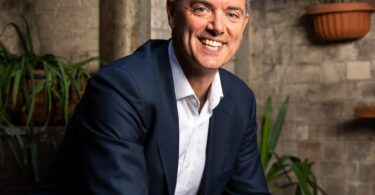 Professional portrait of a smiling man in a navy blazer and white shirt, seated in a plant-filled, brick-walled setting.