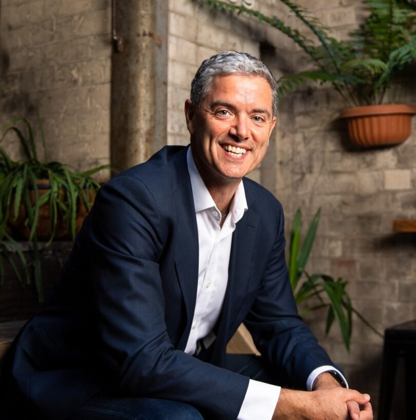 Professional portrait of a smiling man in a navy blazer and white shirt, seated in a plant-filled, brick-walled setting.