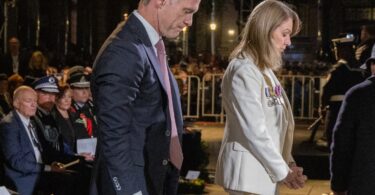 Two formally dressed individuals bow during a ceremony, medals on the woman’s white outfit in the foreground.