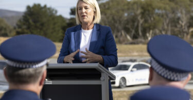 Woman in a blue blazer speaks at an outdoor press briefing behind a lectern, with police officers in the foreground.