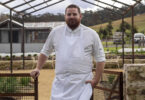 Bearded chef in a white uniform and apron standing by a rusted metal gate outdoors with a skeletal metal roof frame overhead.