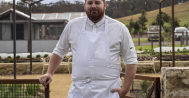 Bearded chef in a white uniform and apron standing by a rusted metal gate outdoors with a skeletal metal roof frame overhead.