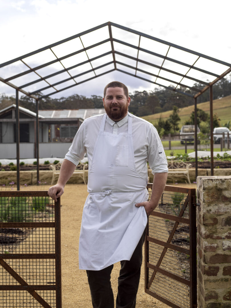 Bearded chef in a white uniform and apron standing by a rusted metal gate outdoors with a skeletal metal roof frame overhead.