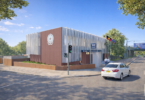 Modern police station with brick and metal panels, blue 'Police' sign, and flagpoles; a white car on the street nearby.