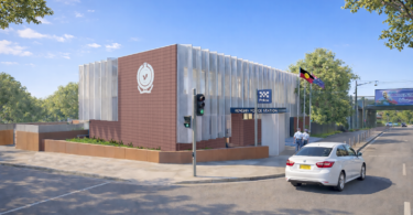 Modern police station with brick and metal panels, blue 'Police' sign, and flagpoles; a white car on the street nearby.