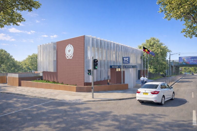 Modern police station with brick and metal panels, blue 'Police' sign, and flagpoles; a white car on the street nearby.