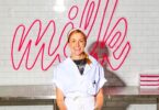 Smiling woman in a white baker's uniform stands at a stainless steel prep counter with a pink neon 'Milk' sign behind her.