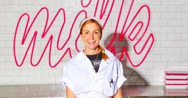 Smiling woman in a white baker's uniform stands at a stainless steel prep counter with a pink neon 'Milk' sign behind her.