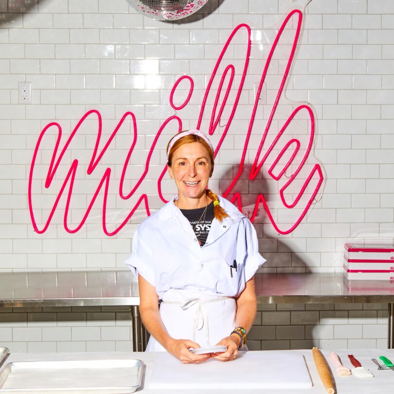 Smiling woman in a white baker's uniform stands at a stainless steel prep counter with a pink neon 'Milk' sign behind her.