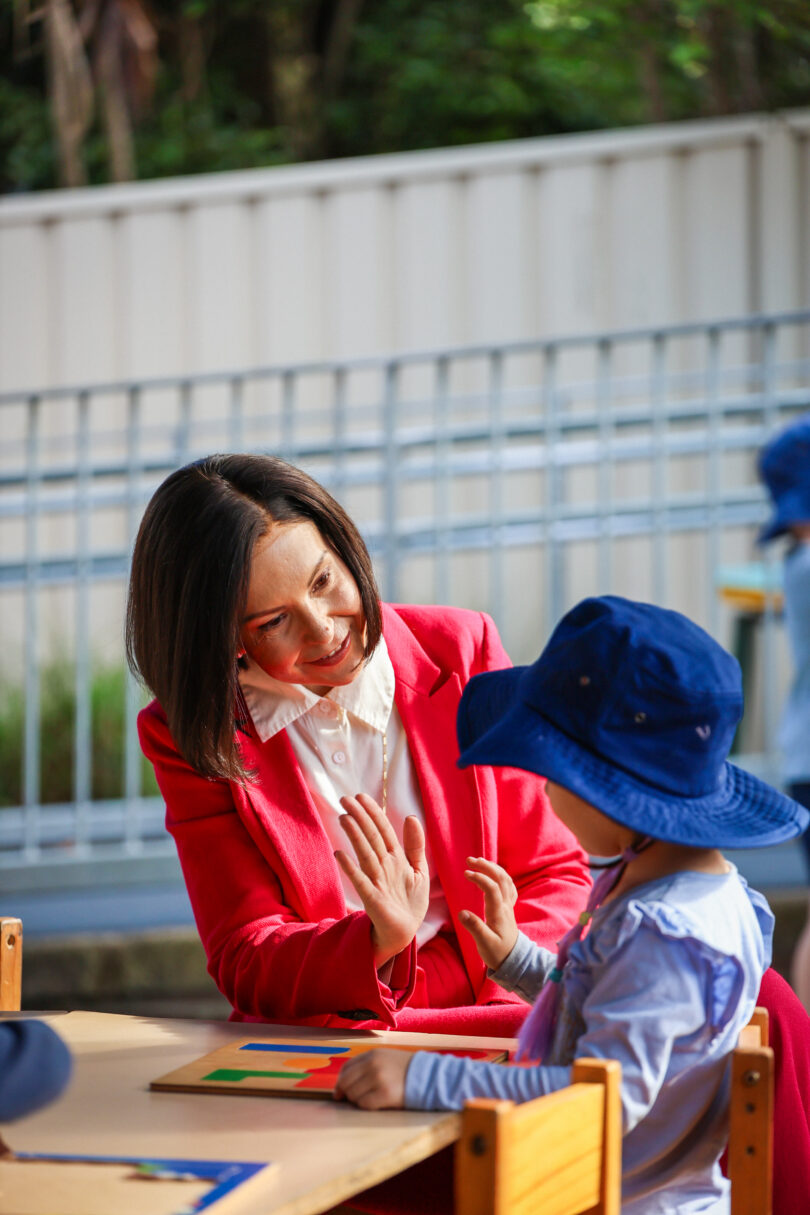 Woman in a red blazer high-fiving a child across a puzzle on a table outdoors