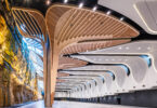 Interior of a modern exhibition hall with sculptural curved wooden arches spanning overhead and a rock cliff wall on the left.