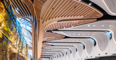 Interior of a modern exhibition hall with sculptural curved wooden arches spanning overhead and a rock cliff wall on the left.