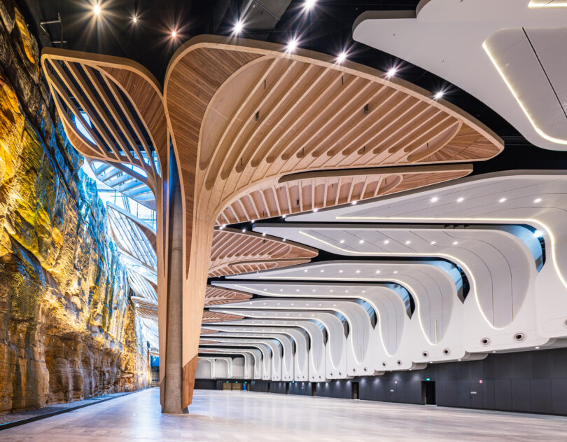 Interior of a modern exhibition hall with sculptural curved wooden arches spanning overhead and a rock cliff wall on the left.