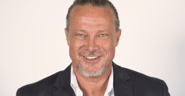 Professional head-and-shoulders portrait of a smiling man in a dark blazer against a light background.