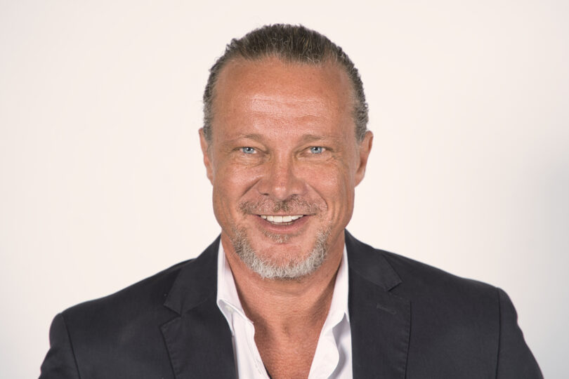 Professional head-and-shoulders portrait of a smiling man in a dark blazer against a light background.