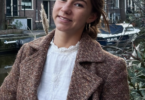 Smiling woman in a brown textured coat standing near a canal with brick buildings in the background.
