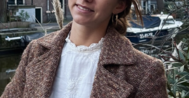 Smiling woman in a brown textured coat standing near a canal with brick buildings in the background.