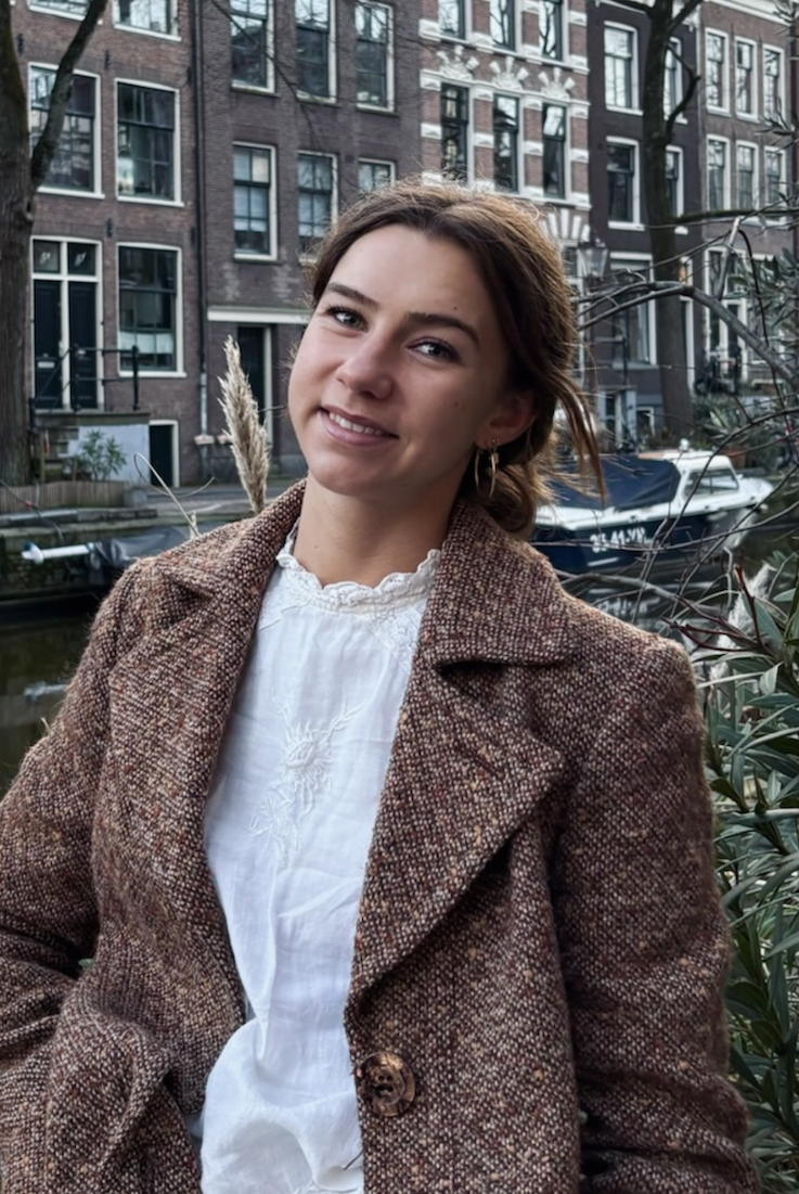 Smiling woman in a brown textured coat standing near a canal with brick buildings in the background.
