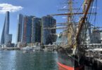 Historic tall ship with rigging docked at a modern city harbor against blue sky and glass skyscrapers behind it.