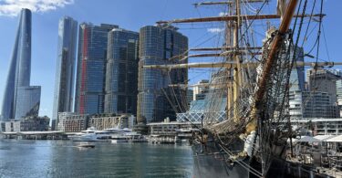 Historic tall ship with rigging docked at a modern city harbor against blue sky and glass skyscrapers behind it.