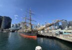 Historic tall ship moored at a harbor beside a modern city skyline under a bright blue sky, water reflecting the hull and rigging.