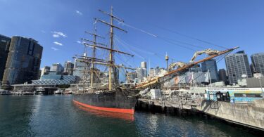 Historic tall ship moored at a harbor beside a modern city skyline under a bright blue sky, water reflecting the hull and rigging.