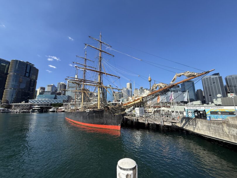 Historic tall ship moored at a harbor beside a modern city skyline under a bright blue sky, water reflecting the hull and rigging.