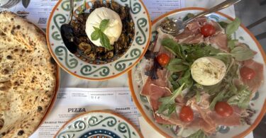 Overhead view of a rustic Italian meal: crusty bread, a bowl of roasted eggplant topped with burrata and fresh basil, and a plate of prosciutto with arugula, burrata, and cherry tomatoes.