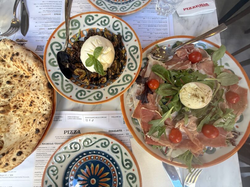 Overhead view of a rustic Italian meal: crusty bread, a bowl of roasted eggplant topped with burrata and fresh basil, and a plate of prosciutto with arugula, burrata, and cherry tomatoes.