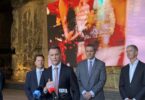 Politician at a podium speaking to press, with reporters' microphones in front and three suited colleagues behind him. Large colorful screen in the background.