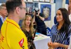 Man in a yellow sports jersey speaks with a woman in a blue striped blouse during an indoor event, with a photographer in the background.