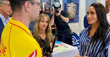Man in a yellow sports jersey speaks with a woman in a blue striped blouse during an indoor event, with a photographer in the background.