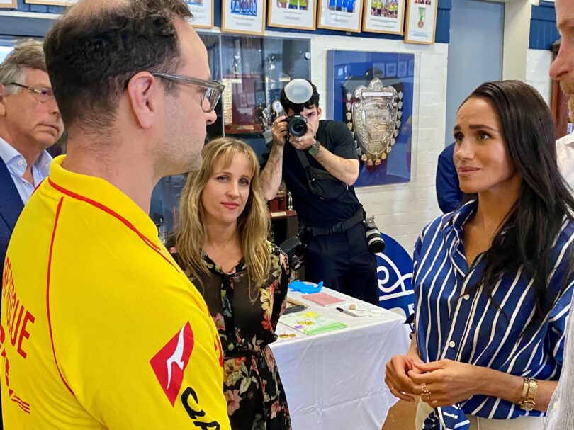 Man in a yellow sports jersey speaks with a woman in a blue striped blouse during an indoor event, with a photographer in the background.