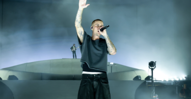 Lead singer on stage, microphone in hand, one arm raised, surrounded by fog and blue lighting.
