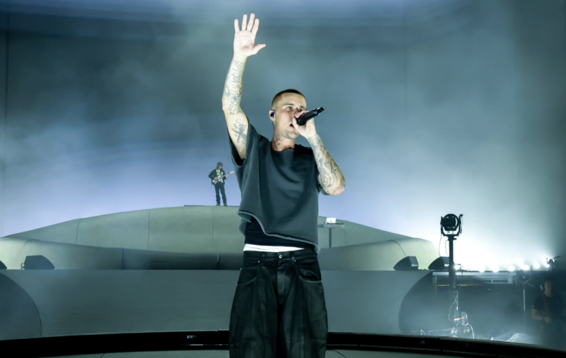Lead singer on stage, microphone in hand, one arm raised, surrounded by fog and blue lighting.